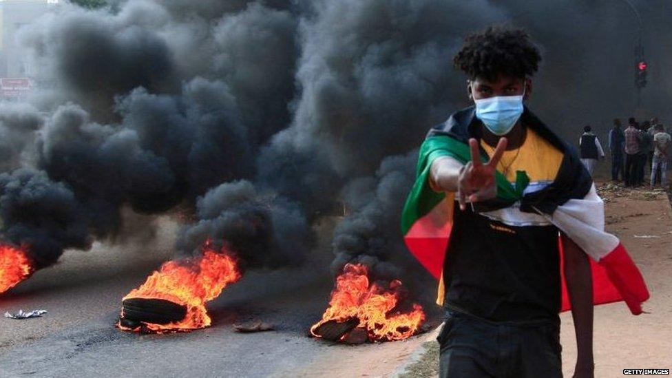 A young boy standing in front of burning tyres with a Sudanese flag draped over him