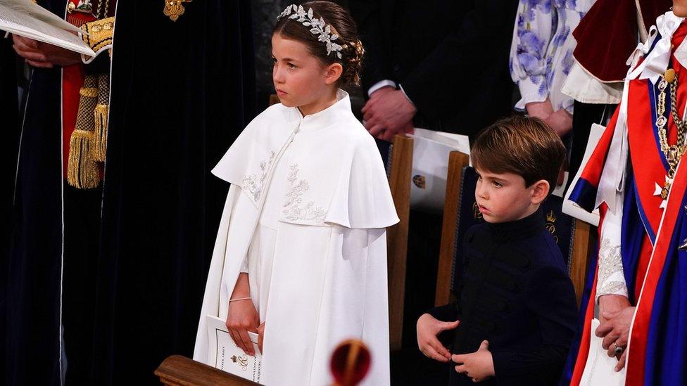 Princess Charlotte is wearing a white cape with embroidery and silver headpiece. Prince Louis is wearing a dark blue leather tunic