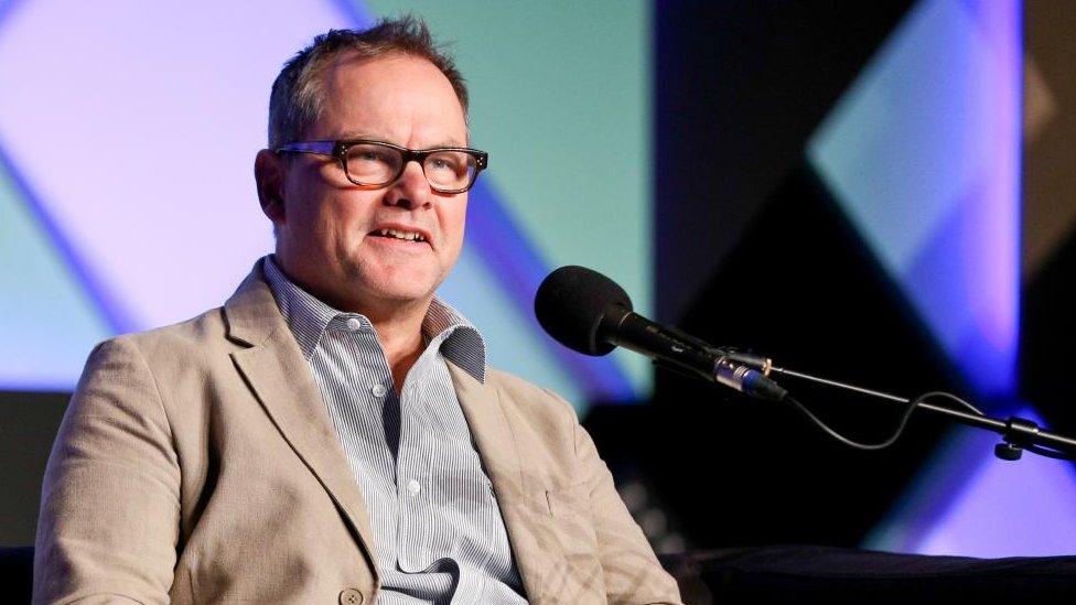 Jack Dee, wearing striped shirt and brown jacket sits behind a microphone