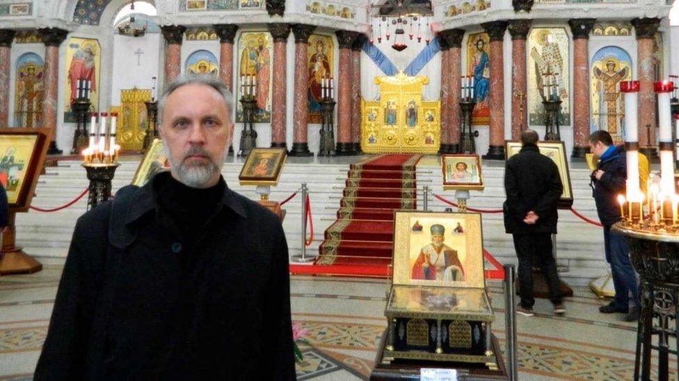 Father Ioann stands in front of Orthodox Church icons.