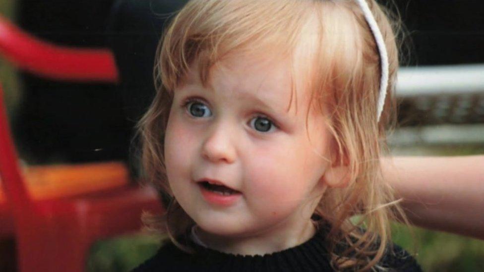 A little girl with strawberry blonde hair wears a head band and a happy expression