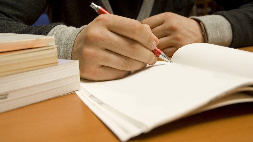 A student is writing in a copy book next to a pile of books.