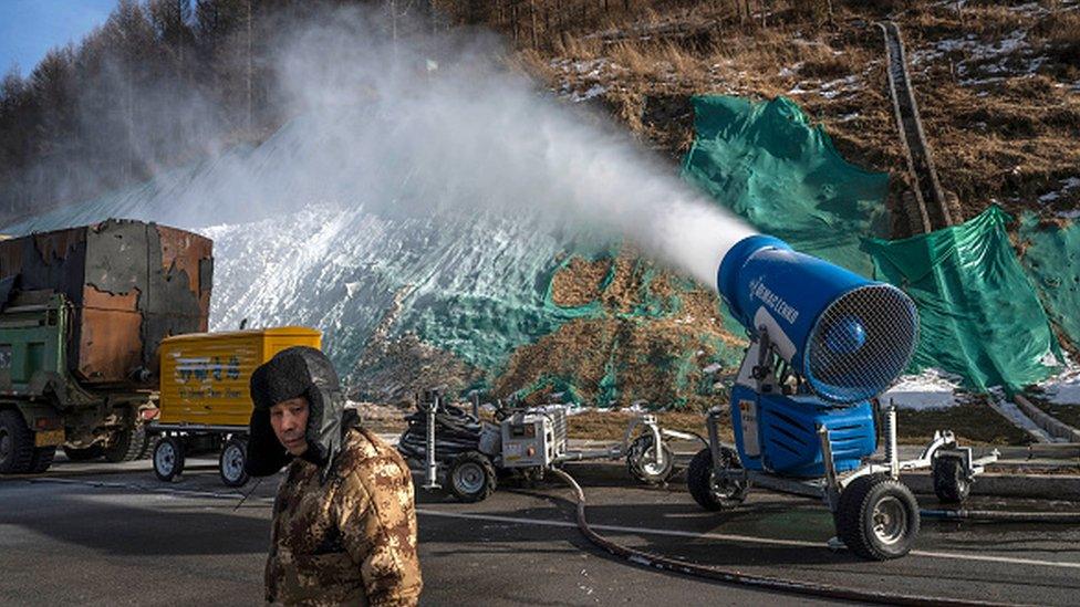 A workers stands next to a snow machine that is making artificial snow