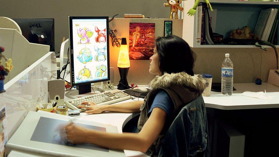 An artist using a drawing tablet on a computer, sat in an office. Her desk has a lava lamp as well as a screen on which six Neopets are visible.