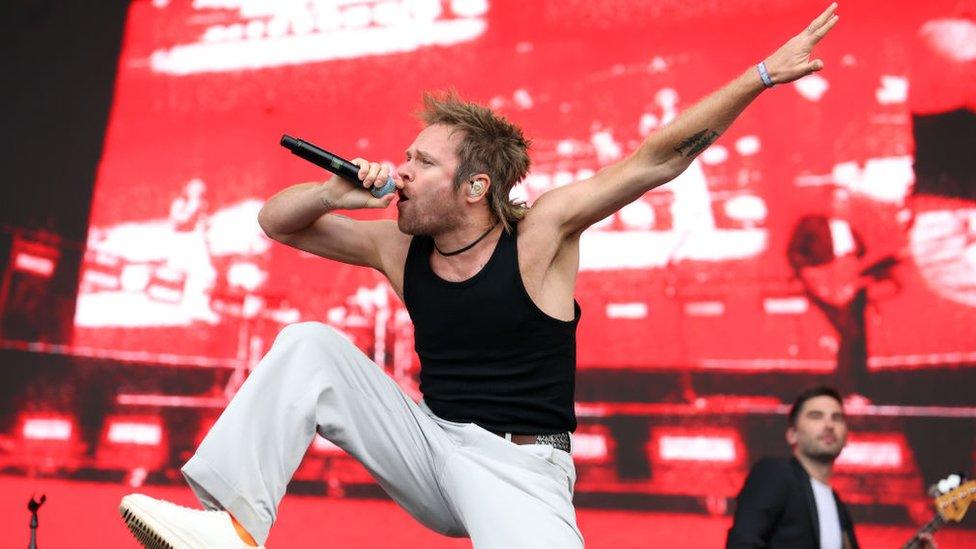 Rou Reynolds of Enter Shikari on stage during day one of Reading Festival 2023. Rou, a white man in his 30s, has light brown hair cut in a mullet. He wears white trousers and a black tank top. His right knee lifted towards him and his left arm outstretched as he sings into a microphone. The stage behind him is lit red