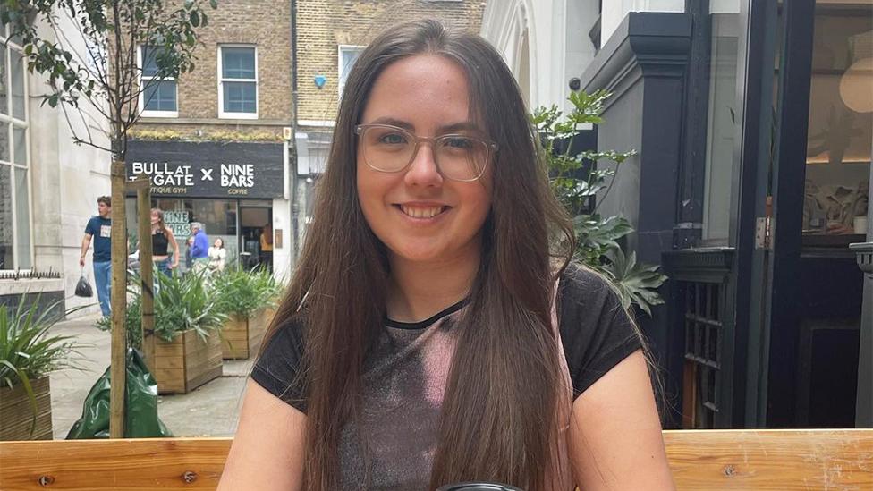 Sophia, wearing glasses, is smiling at the camera. She is sat down surrounded by shops, with plants and trees in the background