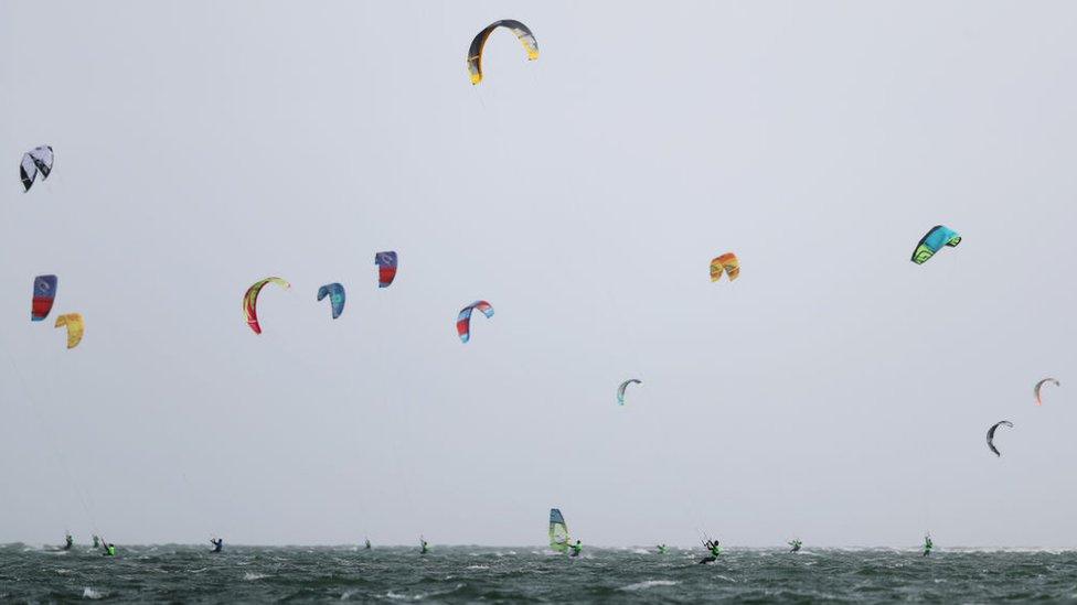 Members of the public are seen kitesurfing and windsurfing at Hayling Island Beach.