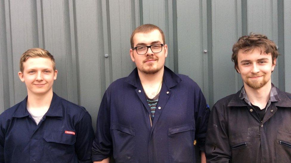 Thomas Allday, Dafydd Hughes and Sam Buffrey stand in a line on front of a corrugated wall in overalls