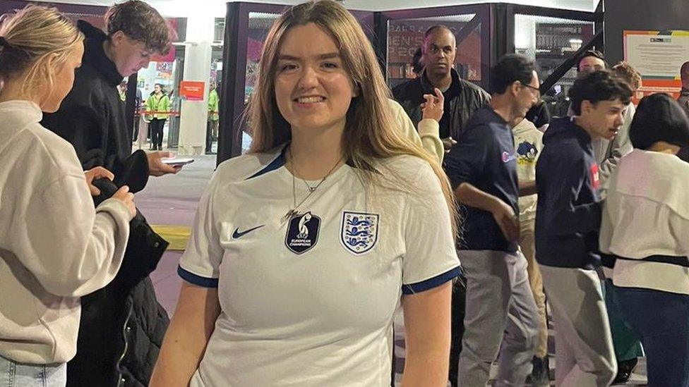 A young white woman with shoulder length brown hair stands in front of a stadium, fans milling about behind her, wearing an England football shirt. Between the Nike swoosh logo and the England Team badge on either side of the chest, in the middle there's a special badge which reads UEFA European Champions. She's smiling and looks relaxed.