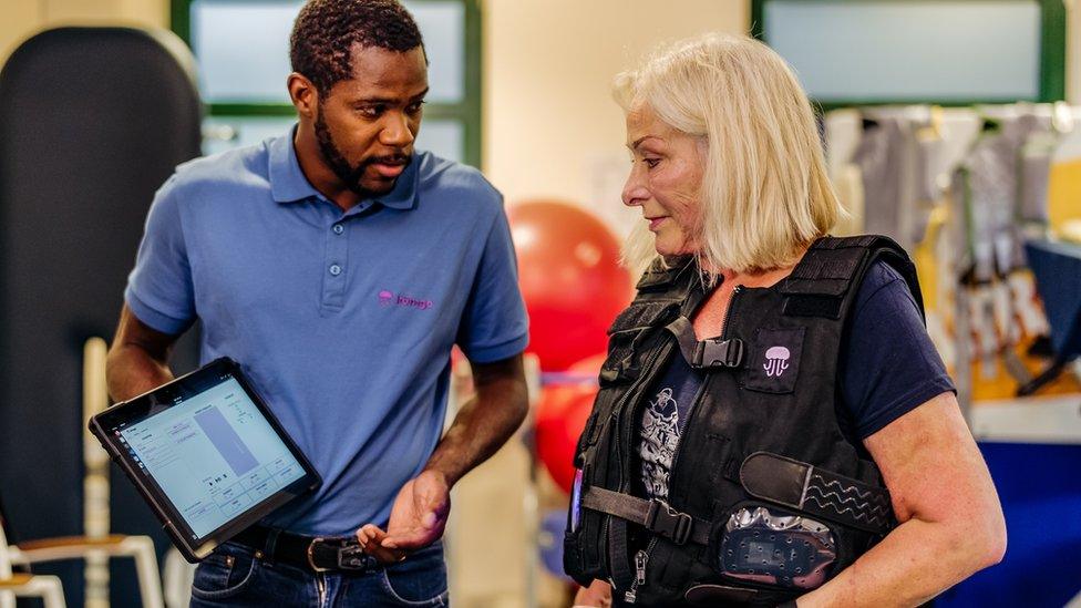 Julie Lloyd in a vest with wiring beside Rudi Gombauld who is holding a pad device controlling the walking device