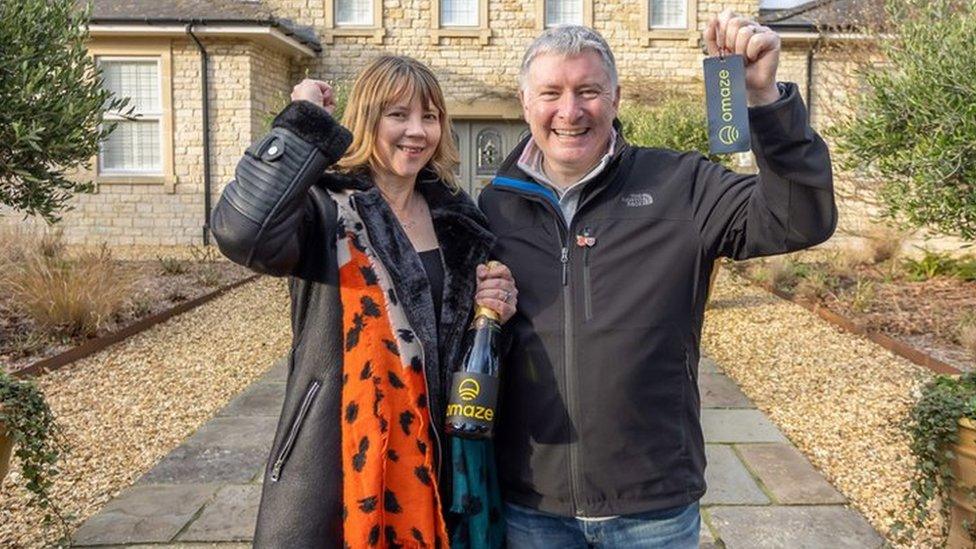 A man and a woman standing in front of a six bedroom house in Somerset, England. Both smiling, the man is holding keys and the woman is holding a black bottle of alcohol