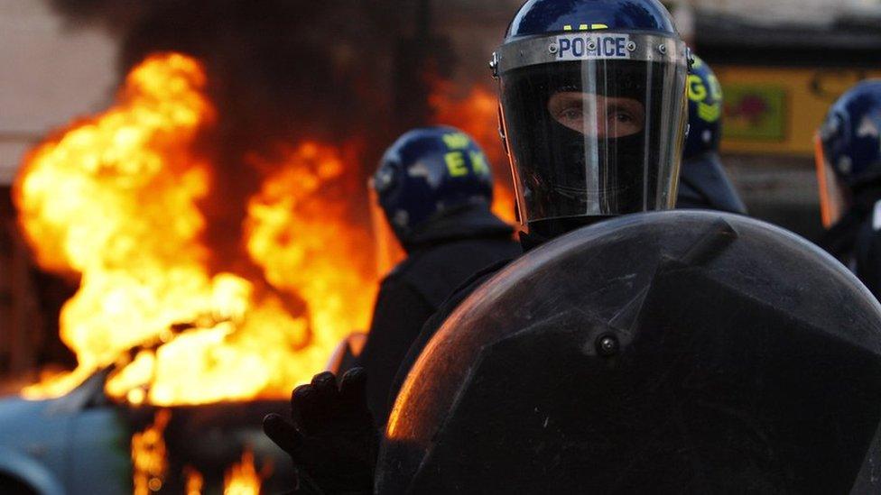 Riot police stand in front of a burning car during riots in London