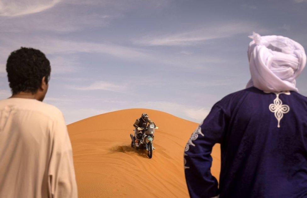 Spectators watch a competitor drive over a dune on a motorbike.