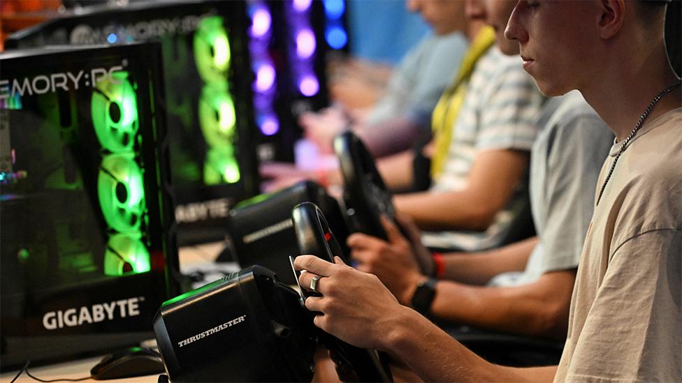 People play a video game at Gamescom 2023 in Cologne, Germany. Three people are pictured with their hands on black steering wheels. There are neon green and purple flashing lights