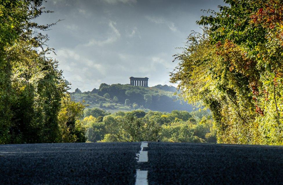 A road between trees with a columned monument on a hill in the background