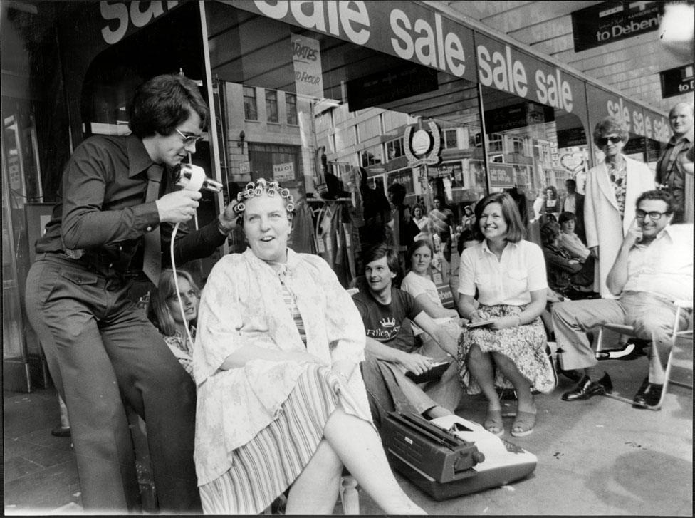 A lady has her blown by a man with a hairdryer as she queues outside a Debenhams store