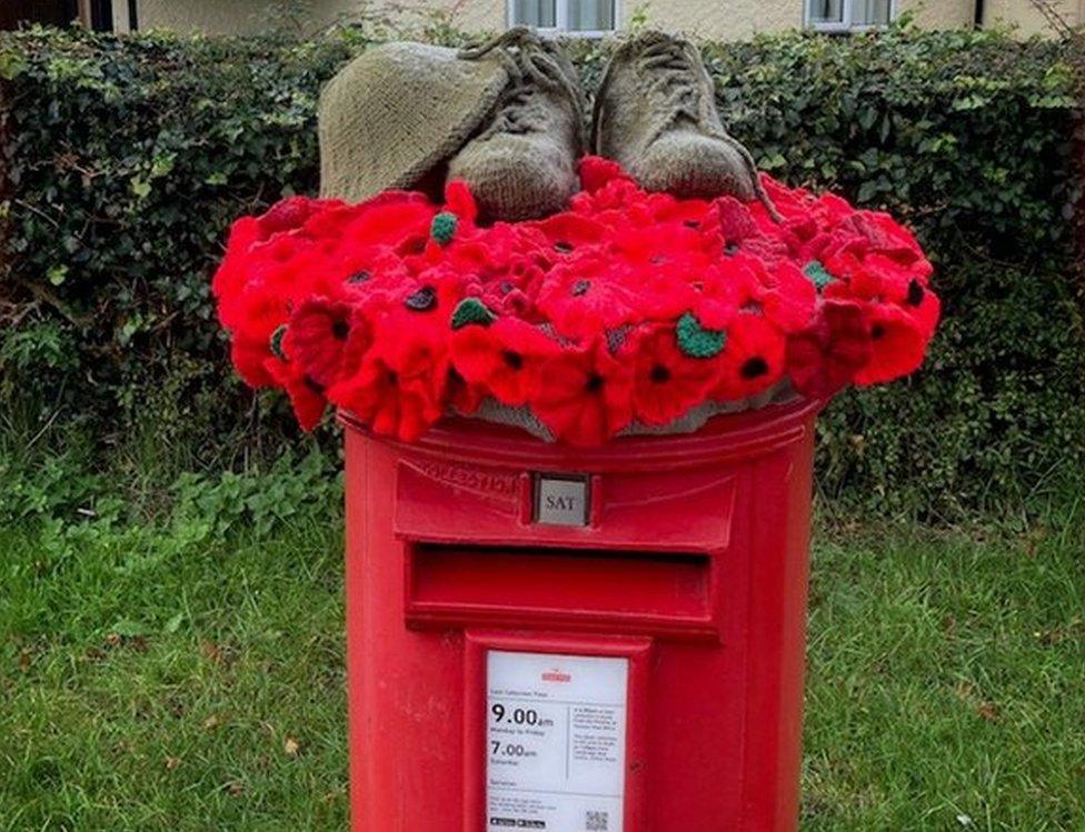 Red post box on grass with a hedge behind. A garland of poppies lies on top, with knitted soldier's shoes and helmet