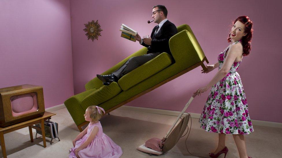 1950s stereotypical housewife uses vacuum cleaner while holding up her husband who's lying on sofa