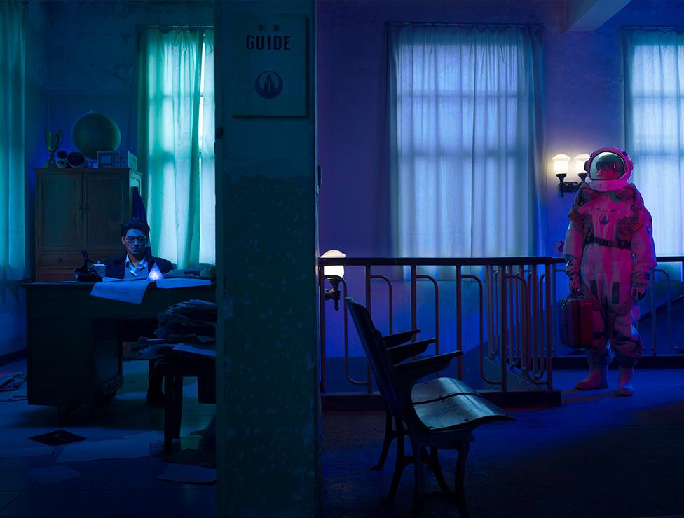 A photo by Cao Fei showing an astronaut looking at a man sitting at a desk