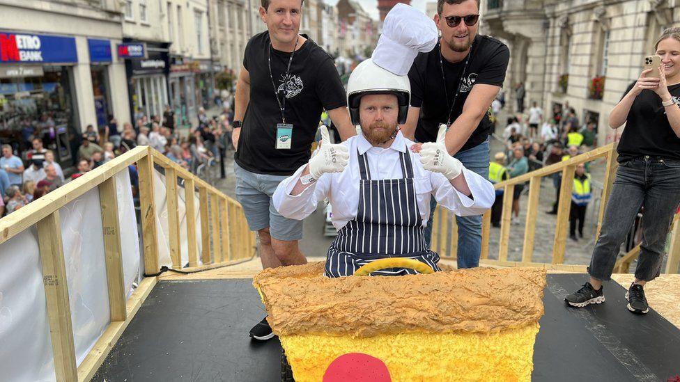 A kart decorated as a pizza slice with the driver dressed as a chef sitting in it and giving a thumbs up to the camera. Two men are pictured behind him ready to push him down a ramp.