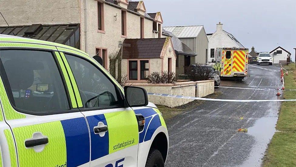 Police car and ambulance and police tape outside row of houses, with puddles on the road.