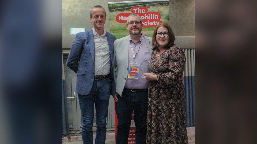 Three people posing for a photo. Two men are wearing suits without ties while a woman with shoulder-length red hair and black glasses is wearing a long dress and holding her award. There is a sign behind them that says "The Haemophilia Society".