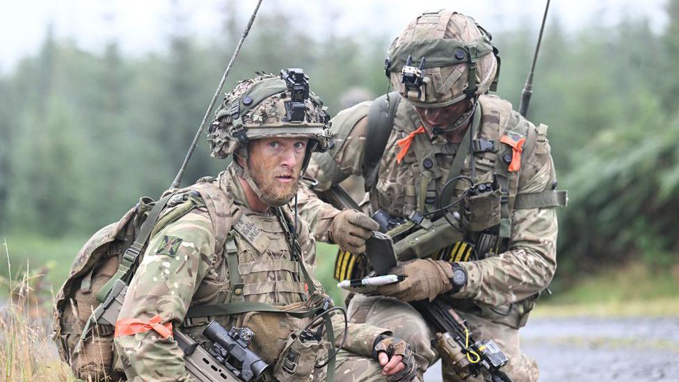 Two soldiers crouched and sitting side by side. They are facing camera and the one on the left is looking straight into the lens.