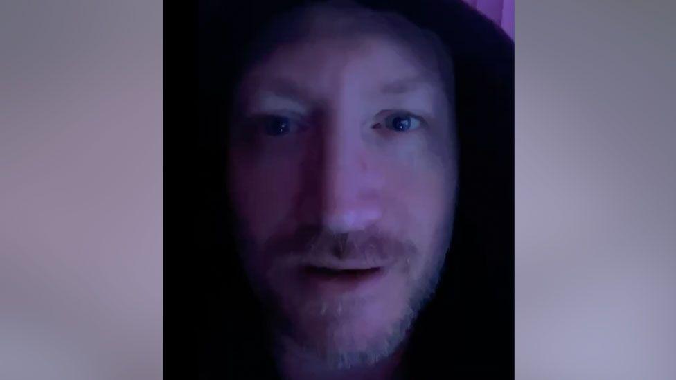 Paul Lipscombe looks straight ahead. He has a moustache and goatee beard. His face is in shadow and he appears to have a hood pulled up on his head