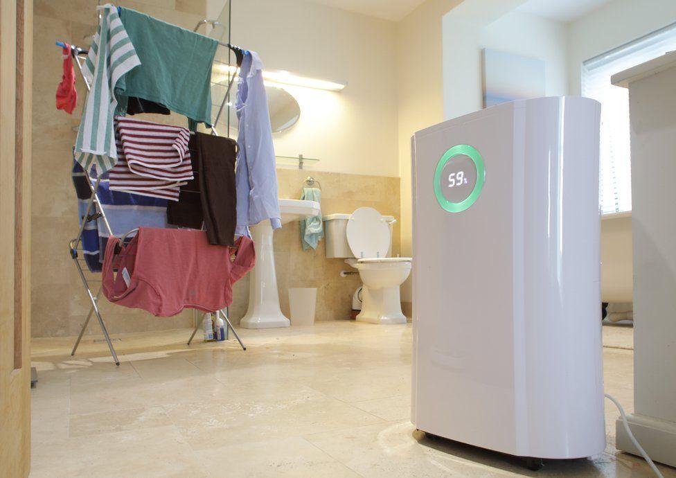 A dehumidifier runs next to a pile of washing which is drying on a clothes rack in a large bathroom. The dehumidifier display reads 59%.