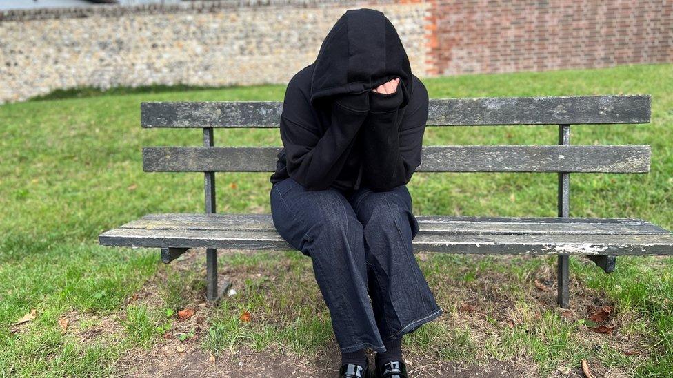 "Amanda" wearing a black hoody with the hood up sat on a bench with her head in her hands