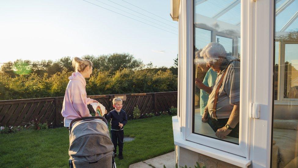 Older people wave through a conservatory to a mother with pram and a child