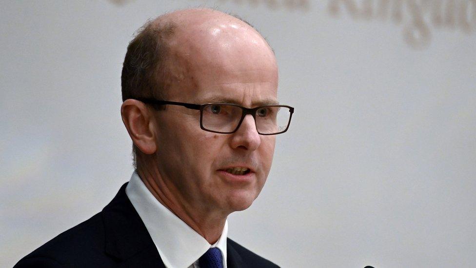 Jeremy Fleming stands against a white background, wearing glasses and a dark suit