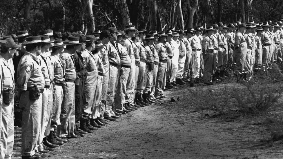 A black and white picture of dozens of police officers stood in a row ready to search a forest