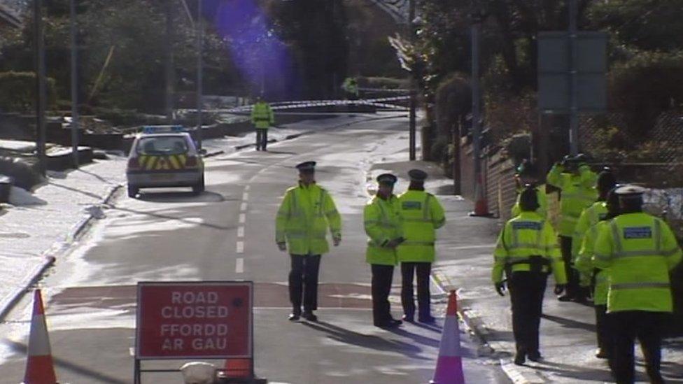 A photo of a cordoned off road. There are eight police officers in high viz jackets, a police car and a sign saying Road Closed