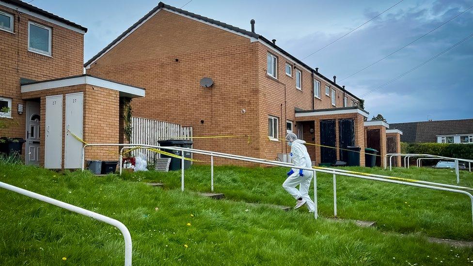 Police officer in a white oversuit walking up the steps towards the entrance of a home that has yellow police warning tape around it