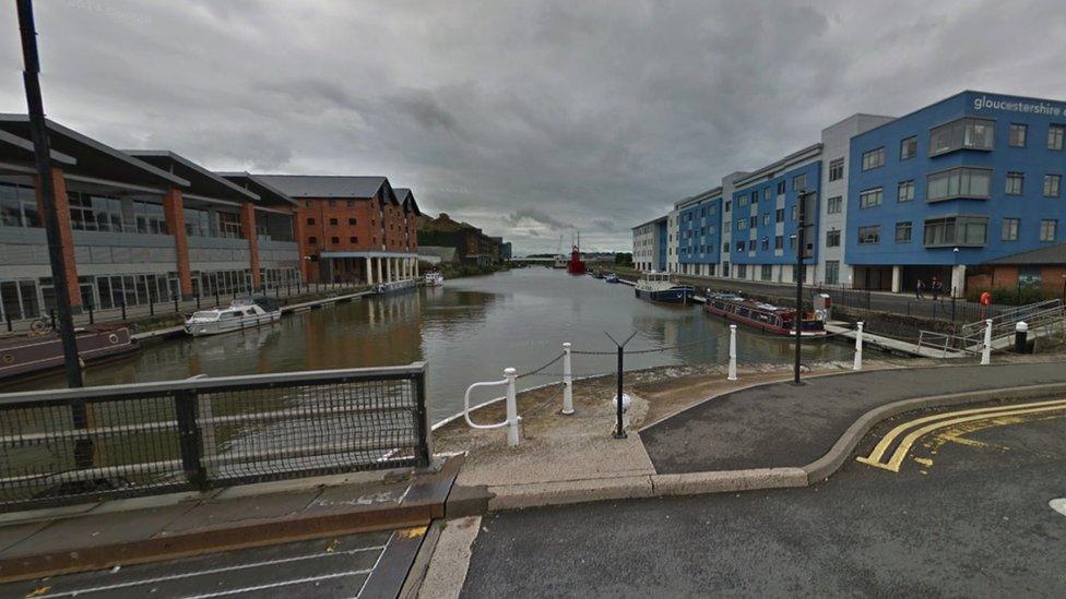 Google maps image of Llanthony Road in Gloucester. The Docks can be seen from the bridge and there are buildings either side of the canal. Gloucestershire College is on the right - it is a blue building with the name written in white letters at the top.