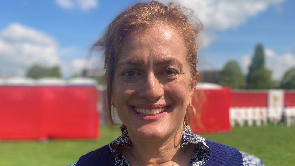 Tasmia Power is standing with grass and red screens in the background. She is smiling and wearing a blue and white patterned blouse with a buttoned up blue waistcoat over it