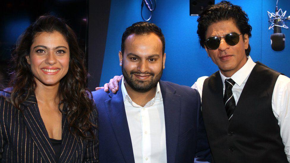 A glamourous woman in a pinstripe suit stands next to a younger man in a navy suit with an open-necked white shirt. To the right of them another man in a white shirt and black waistcoat wears sunglasses and rests his hand on the other man's shoulder. In the background a microphone hangs from the ceiling, telling the viewer the trio are in a studio of some sort.