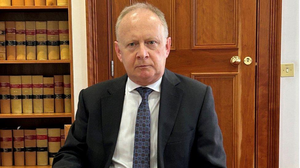 Lord Turnbull, wearing a dark suit and tie, sits in front of a wooden door and a bookcase filled with legal texts