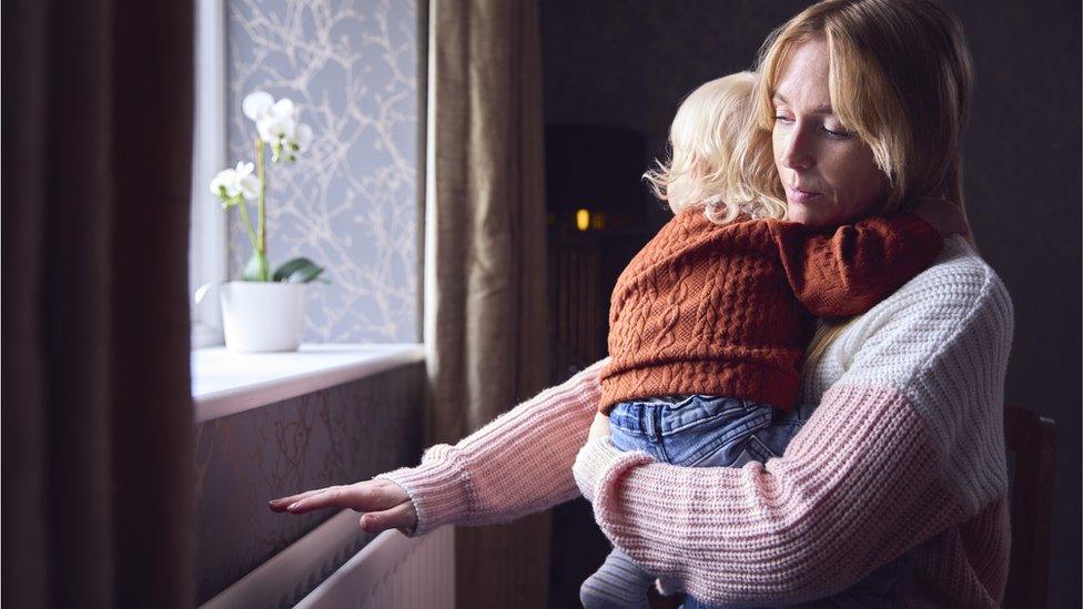 Woman stands near radiator holding a small child