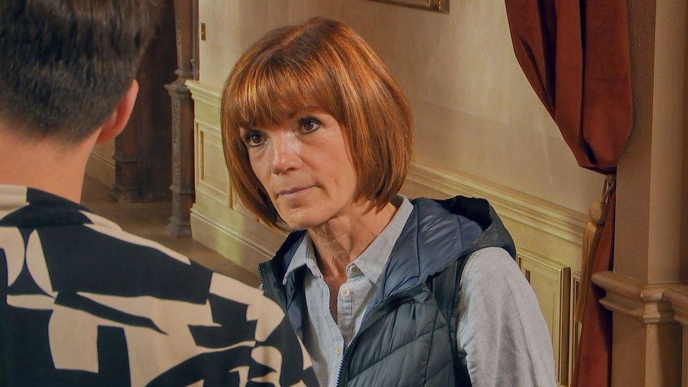 Diane pictured in episode 3 of The Traitors. Diane is a 63-year-old white woman with a ginger bob and brown eyes. She wears a blue gilet over a pale blue shirt and is seen giving suspicious eyes to fellow player Brian who is pictured from the back wearing a patterned black and white shirt. They are inside in the castle hallway which is painted in light colours with dark red drapes and wooden features