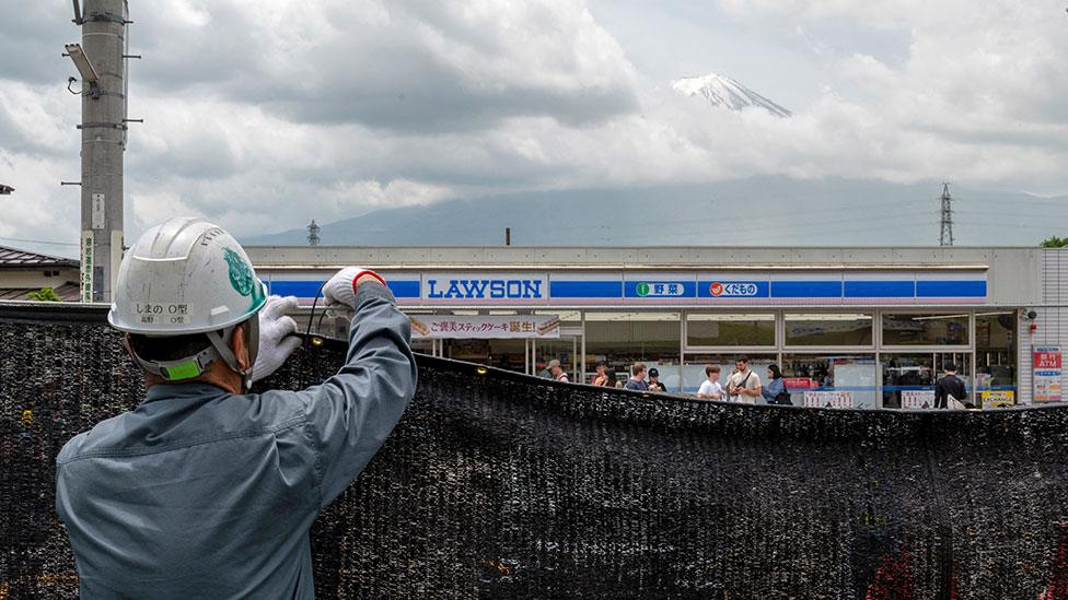 A worker puts up a black barrier in front of a convenience store, blocking a view of Mount Fuji