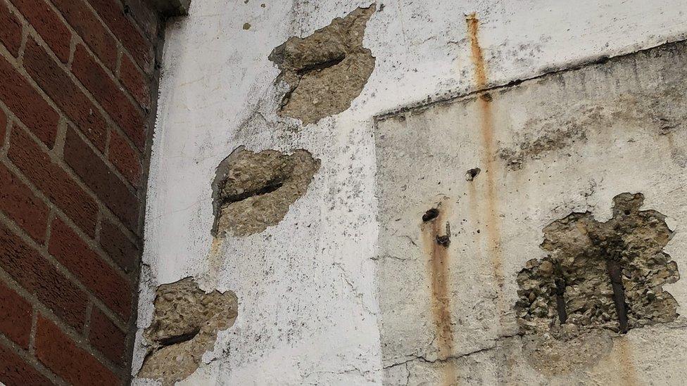 Side of multi-storey car park showing patches where concrete has worn away, rusting the steel bars underneath.