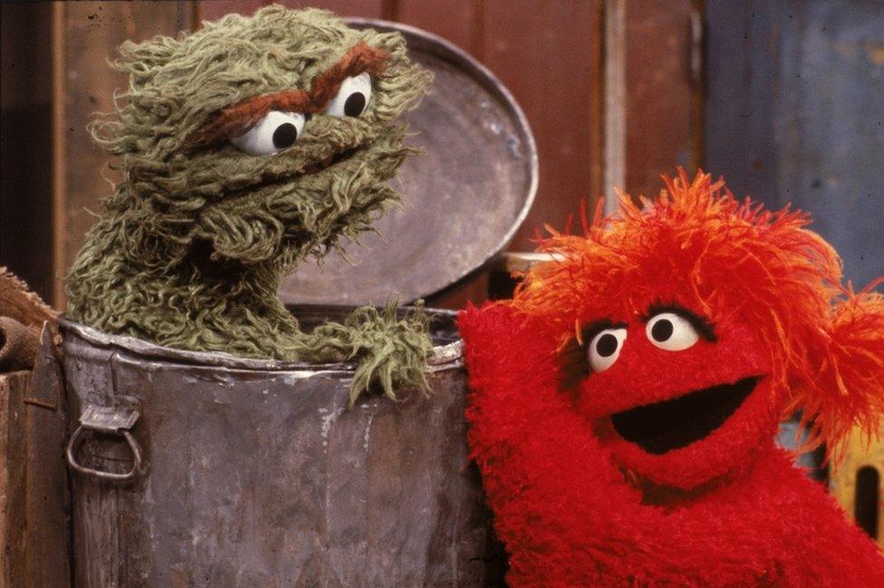 A red muppet visits Oscar the Grouch, pictured inside his garbage can, in a 1980s scene from Sesame Street