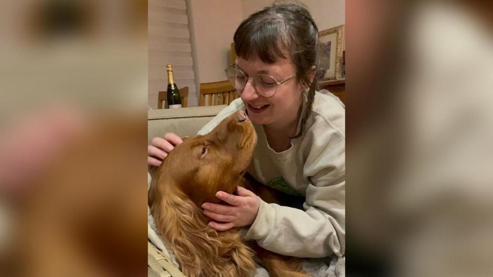 Becky Burke smiles as she hugs Bo Jangles. She has long plaited brown hair and is wearing glasses. A bottle of sparkling wine - possibly champagne - can be seen on a table behind them.
