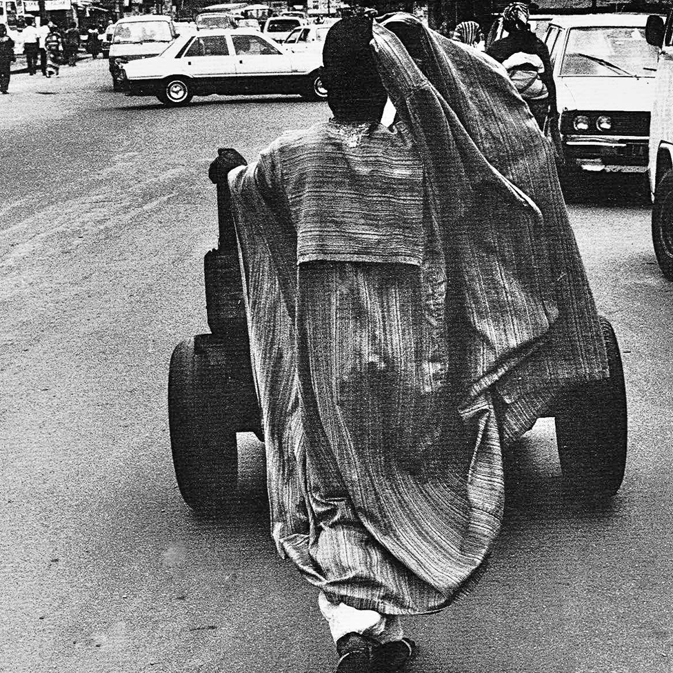 A photo by Sunmi Smart-Cole entitled: "Right Job, Wrong Attire" - 1987 showing a man wearing a boubou pushing a cart