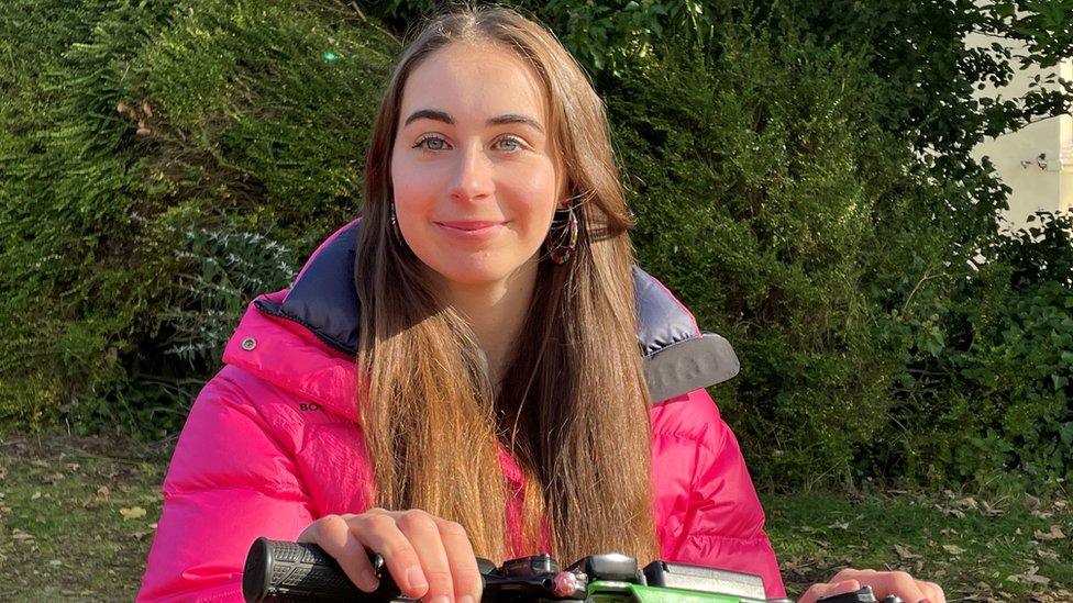 Bethany Handley sitting in her wheelchair in a park. She is a young female wearing a bright pink coat.