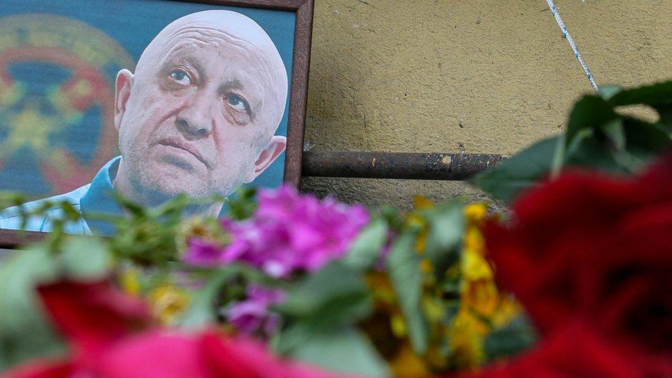 A framed image of Yevgeny Prigozhin is seen with floral tributes in the foreground