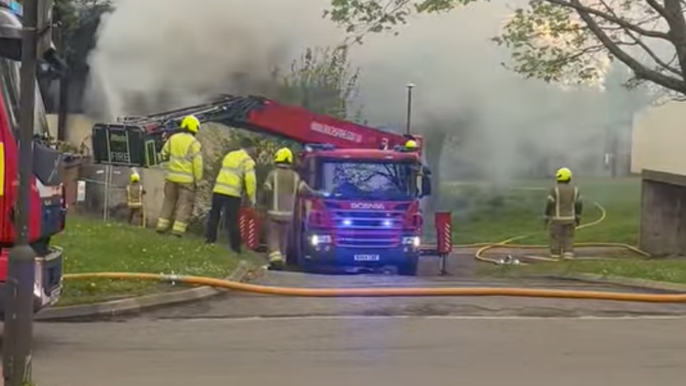 A fire engine and four firefighters. There is a lot of smoke behind the fire engine