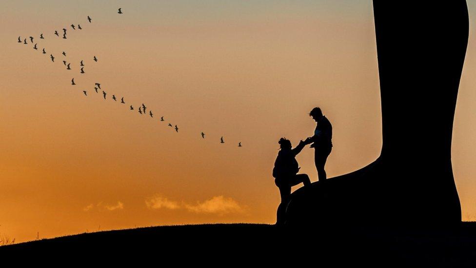 Silhouette of a man helping a woman to climb on the feet of the Angel of the North with birds flying across an orange sky beyond